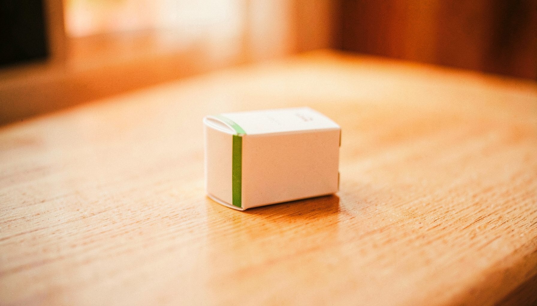 A small white medication box with a subtle green stripe, placed on a light wooden surface with soft natural light.